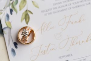 Wedding invitation with gold calligraphy text and wedding rings on floral stationery.