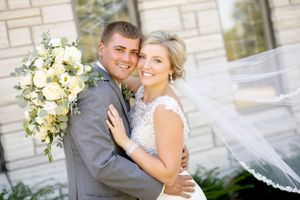A couple poses with a beautiful white floral bouquet against a light stone wall.
