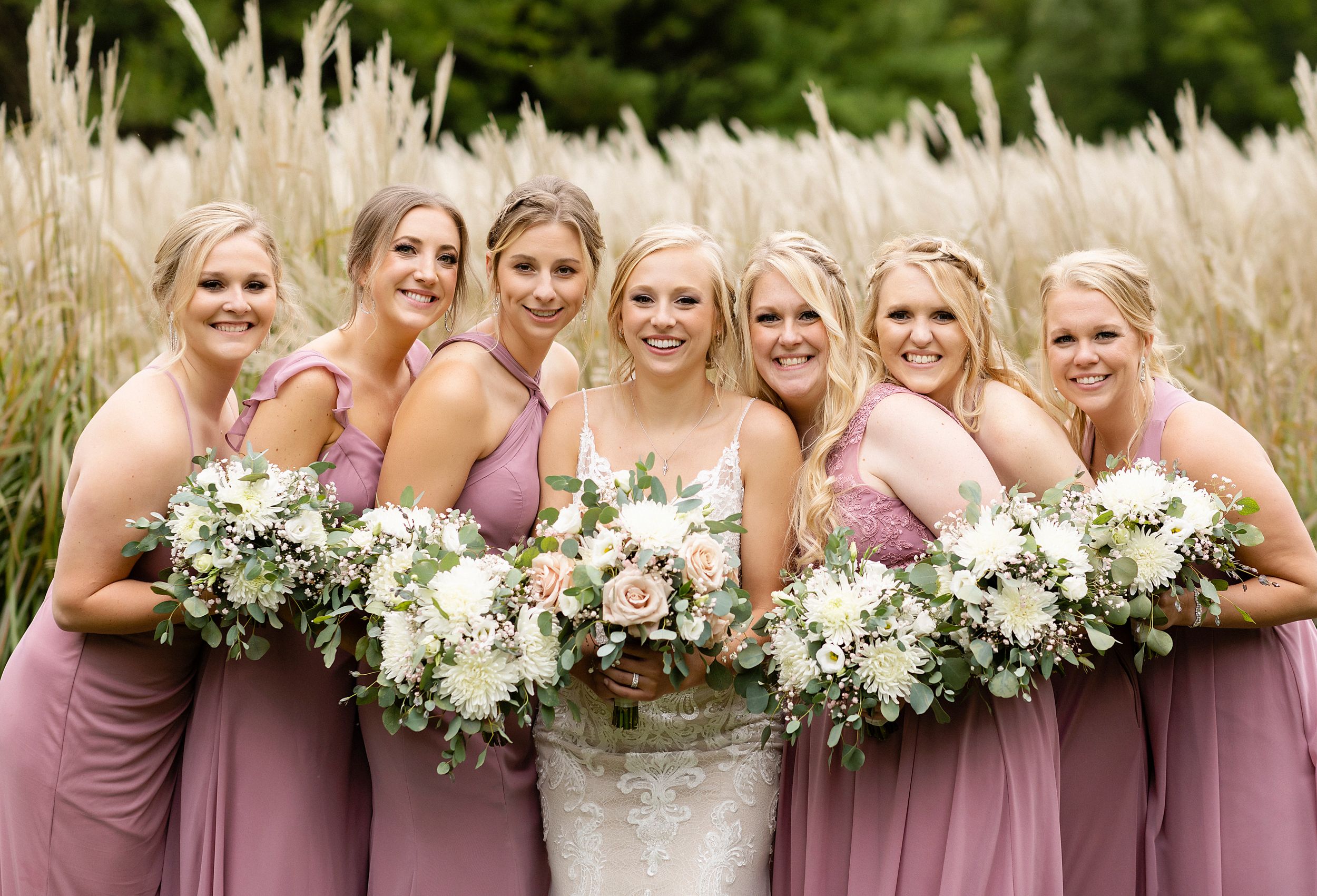Bridesmaids in dusty rose dresses hold white and green bouquets while posing in tall grass.