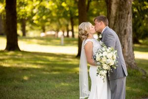 A couple shares an intimate moment in a sun-dappled park with yellow and white bouquet.