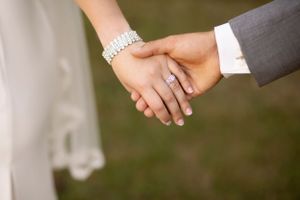 Close-up of newlyweds holding hands, showing wedding rings and bridal bracelet.