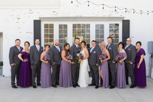 Wedding party in purple and gray attire posed against white building with string lights.