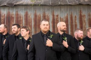 Groomsmen in matching black suits with boutonnieres pose in front of a rustic metal wall.