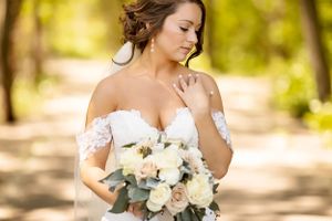 A bride in a white gown holds a beautiful bouquet of roses and eucalyptus in a sunlit forest setting.