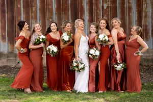 Bridesmaids in matching rust-colored dresses hold bouquets while posing against a rustic backdrop.