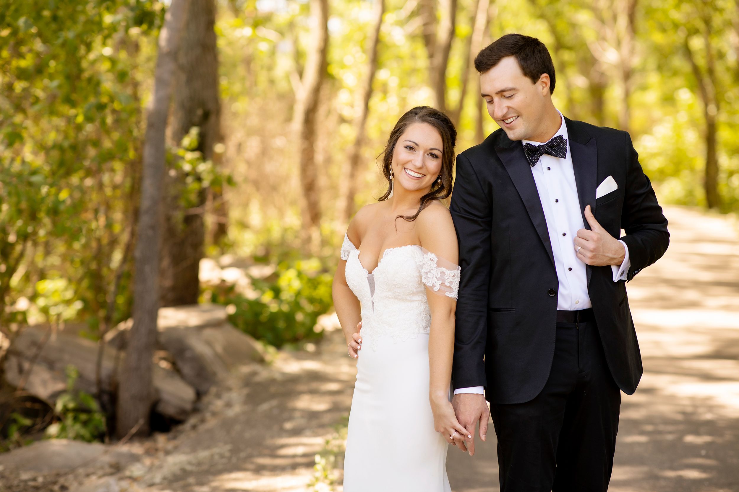 A newlywed couple walks together on a wooded path in formal wedding attire.