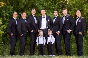 Wedding party in formal attire poses outdoors with two young ring bearers in suspenders.
