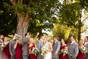 Wedding party in grey suits and red dresses celebrates under tree canopy.