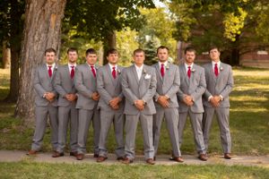 Groomsmen in matching grey suits with red ties line up for formal photo outdoors.