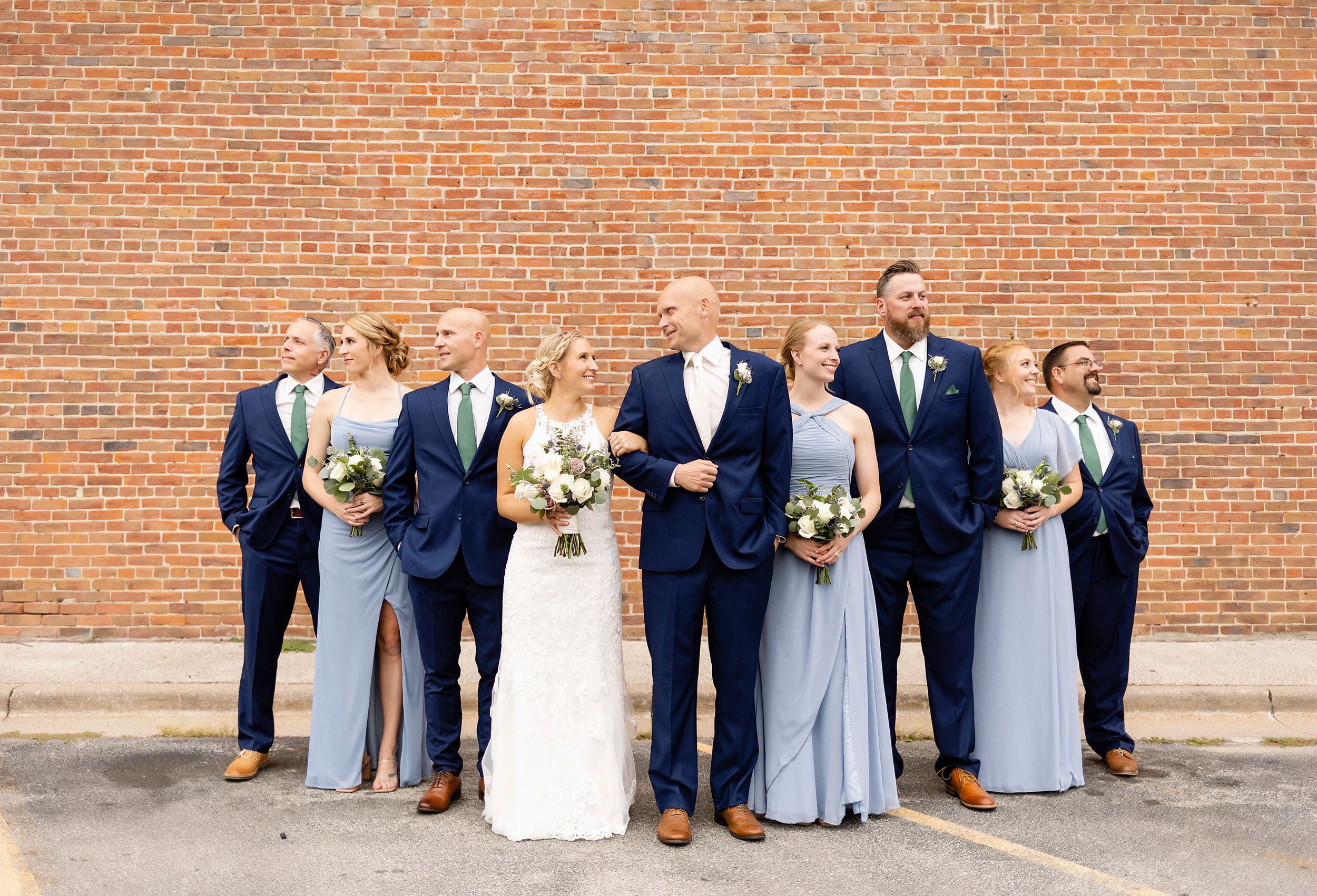 Wedding party in navy suits and light blue dresses poses against brick wall.