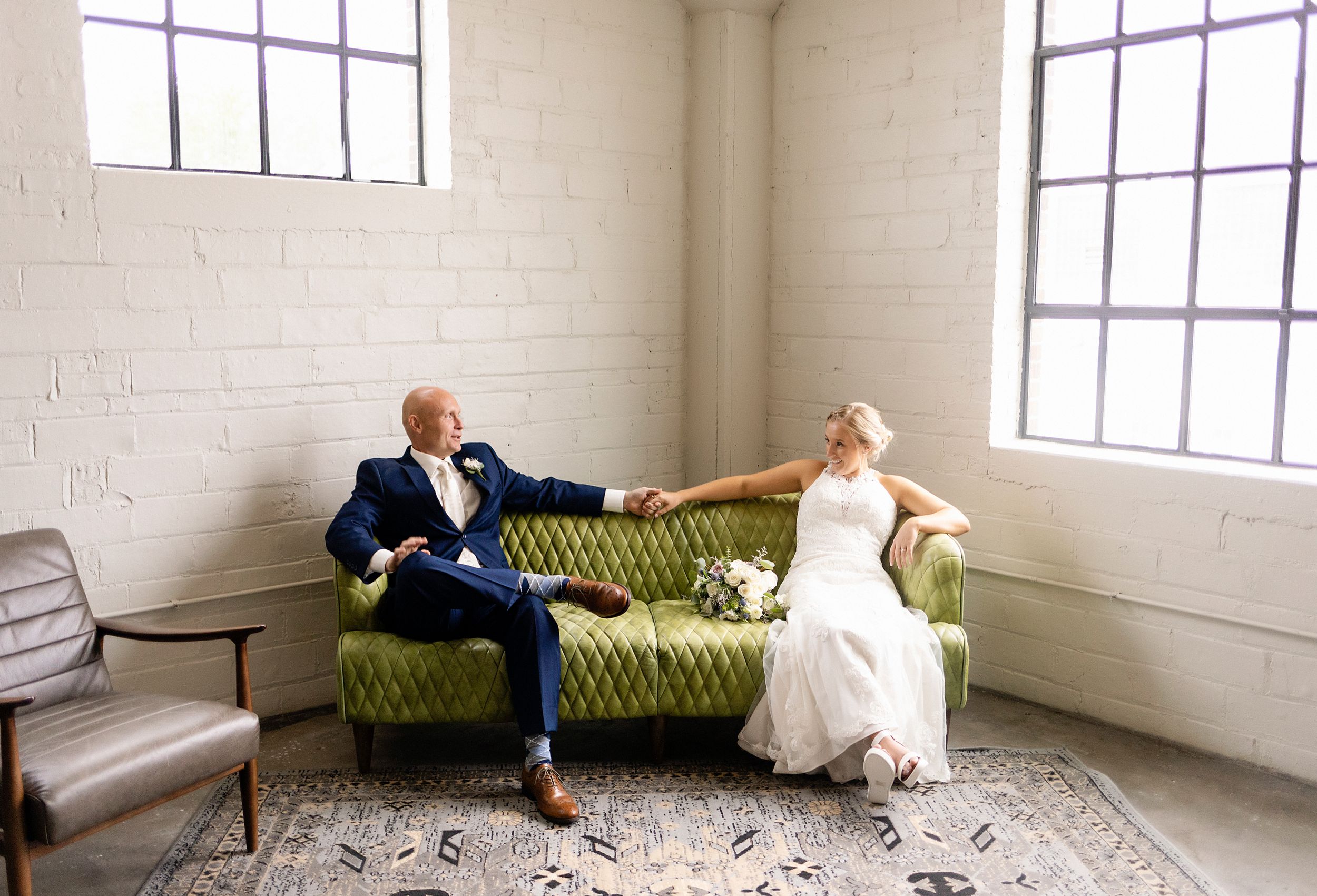 Wedding couple relaxes on vintage green tufted sofa in bright white room.