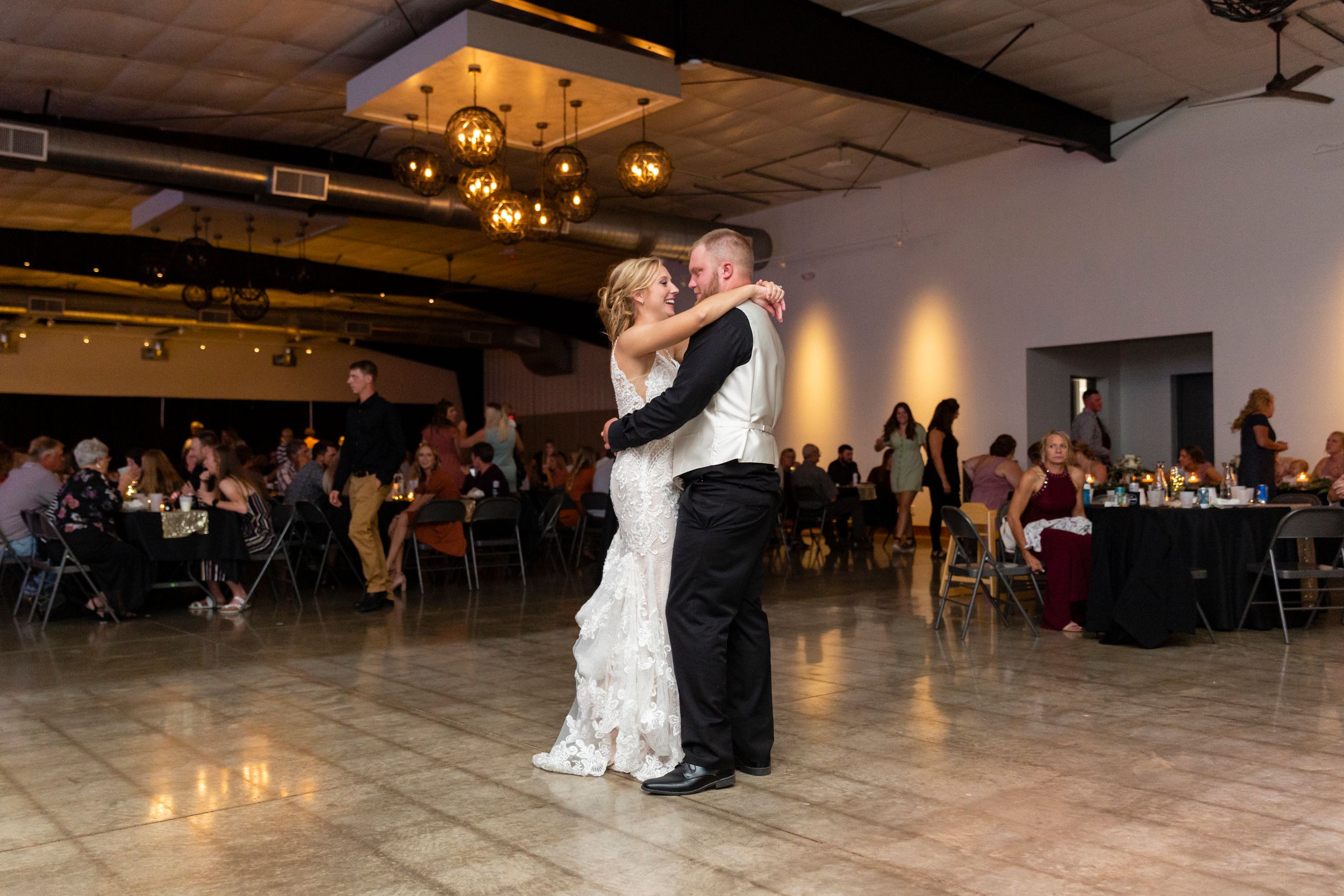 A couple shares their first dance in an elegantly lit reception venue with guests seated in the background.
