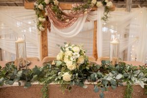 Elegant wedding table decor with white floral centerpiece, draped fabric, and greenery garland against wooden arch.