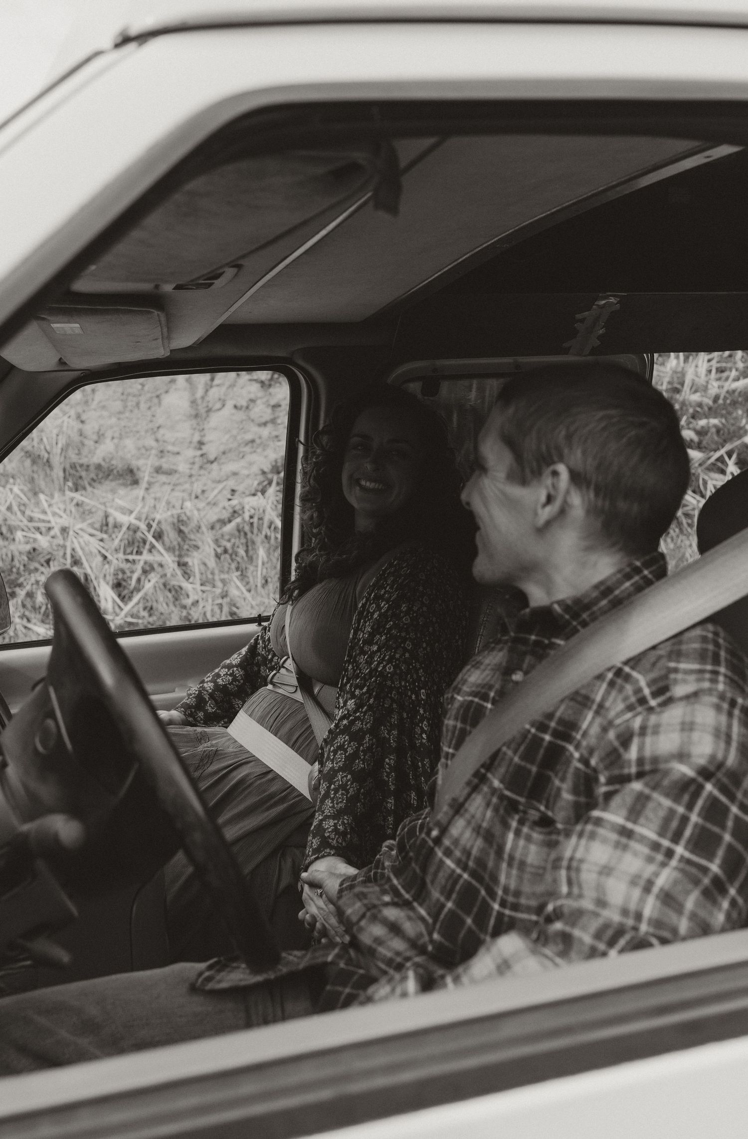 A black and white photo of two people sitting in the front seats of a car, with one person wearing a plaid shirt.