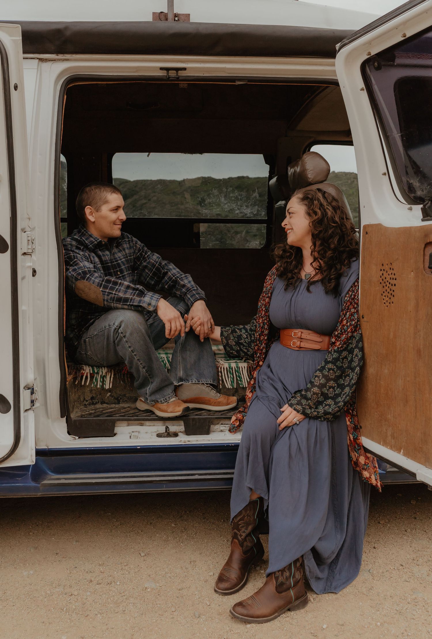 A couple relaxes inside and beside a camper van parked in a scenic mountainous area wearing vintage-inspired outfits.
