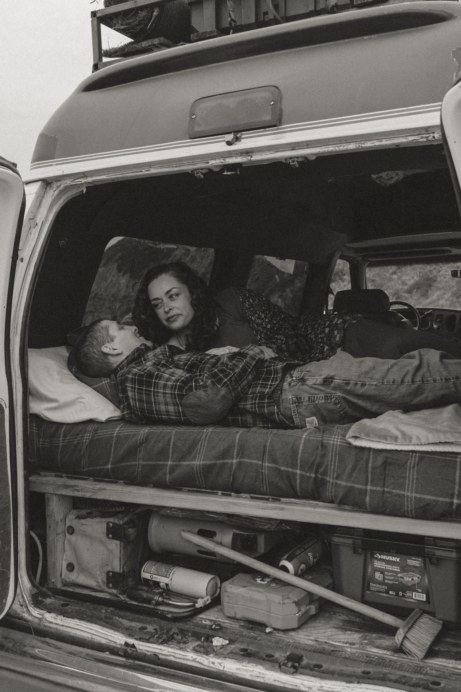A black and white photo of people resting in the back of a van on plaid bedding with camping gear visible below.
