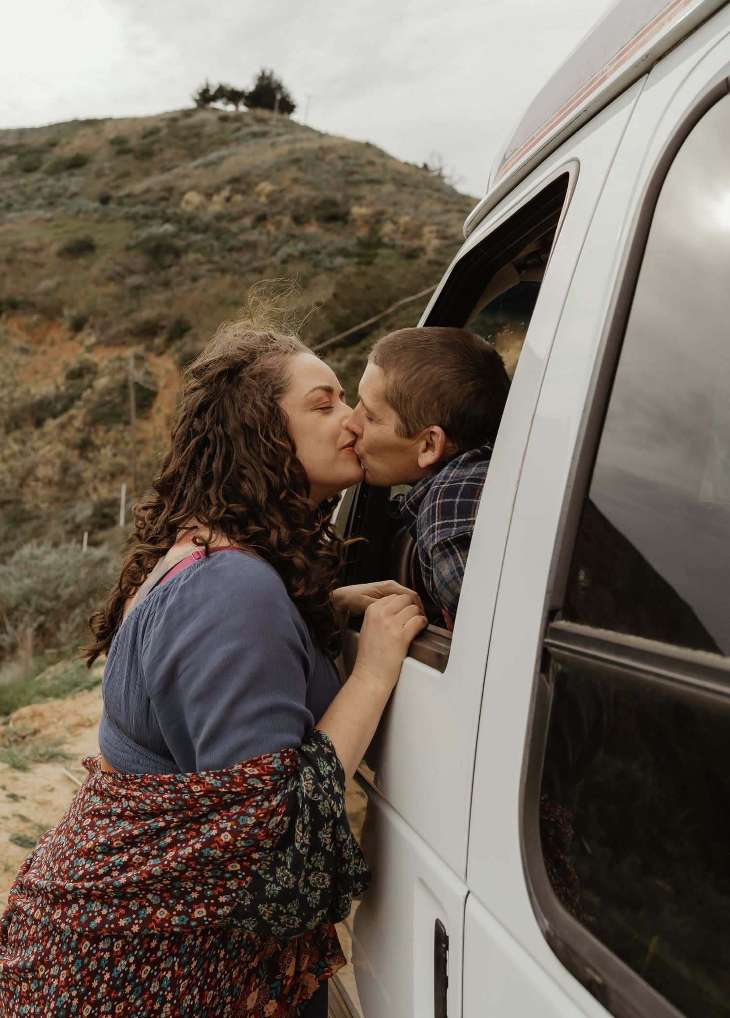 A romantic moment is shared between two people through the window of a parked van in a desert landscape.