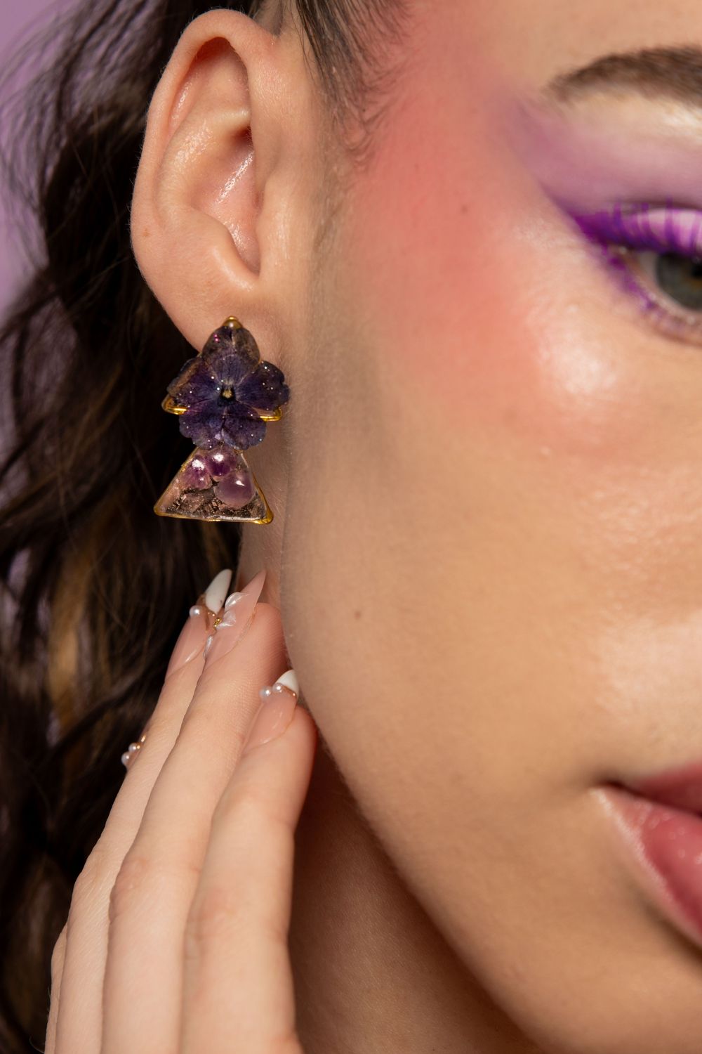 Close-up of woman wearing purple floral triangle drop earrings with matching purple eye makeup and French tip nails.