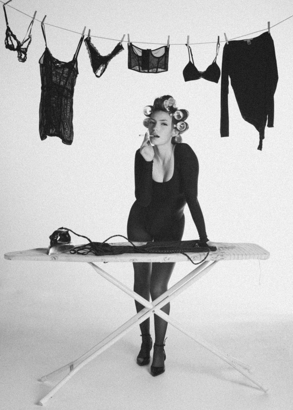 Woman in black bodysuit with hair rollers posing at ironing board, black and white fashion photo.
