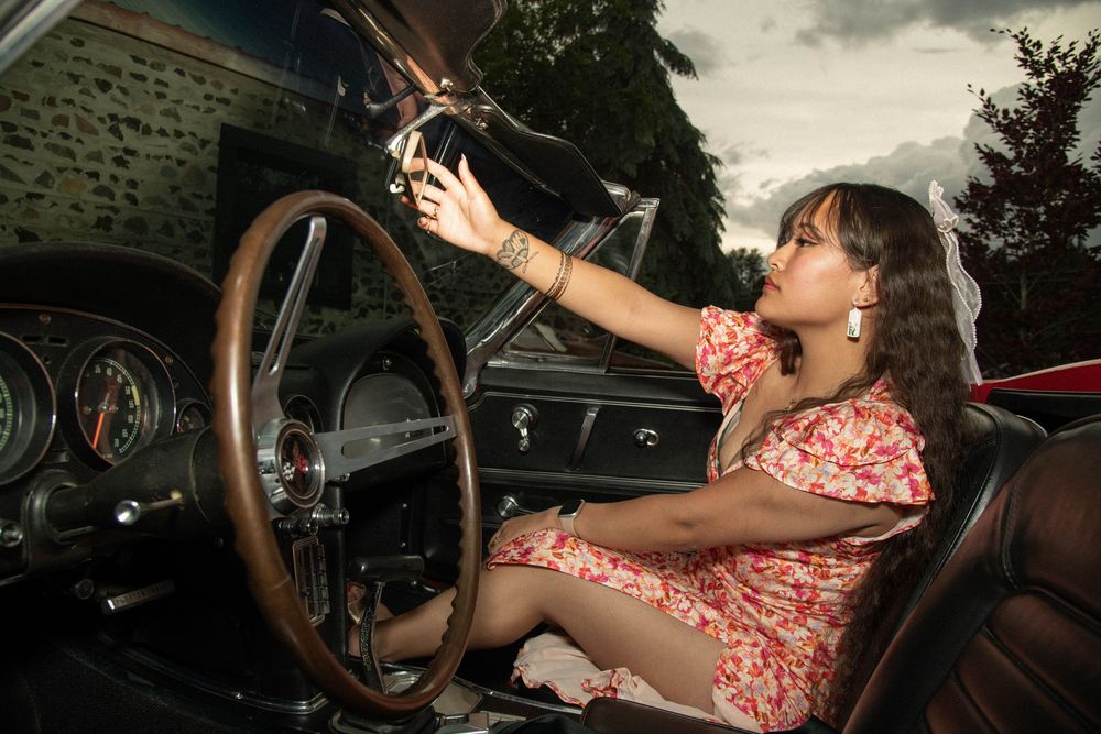 Young woman in floral dress reaching for radio inside a classic vintage car interior.