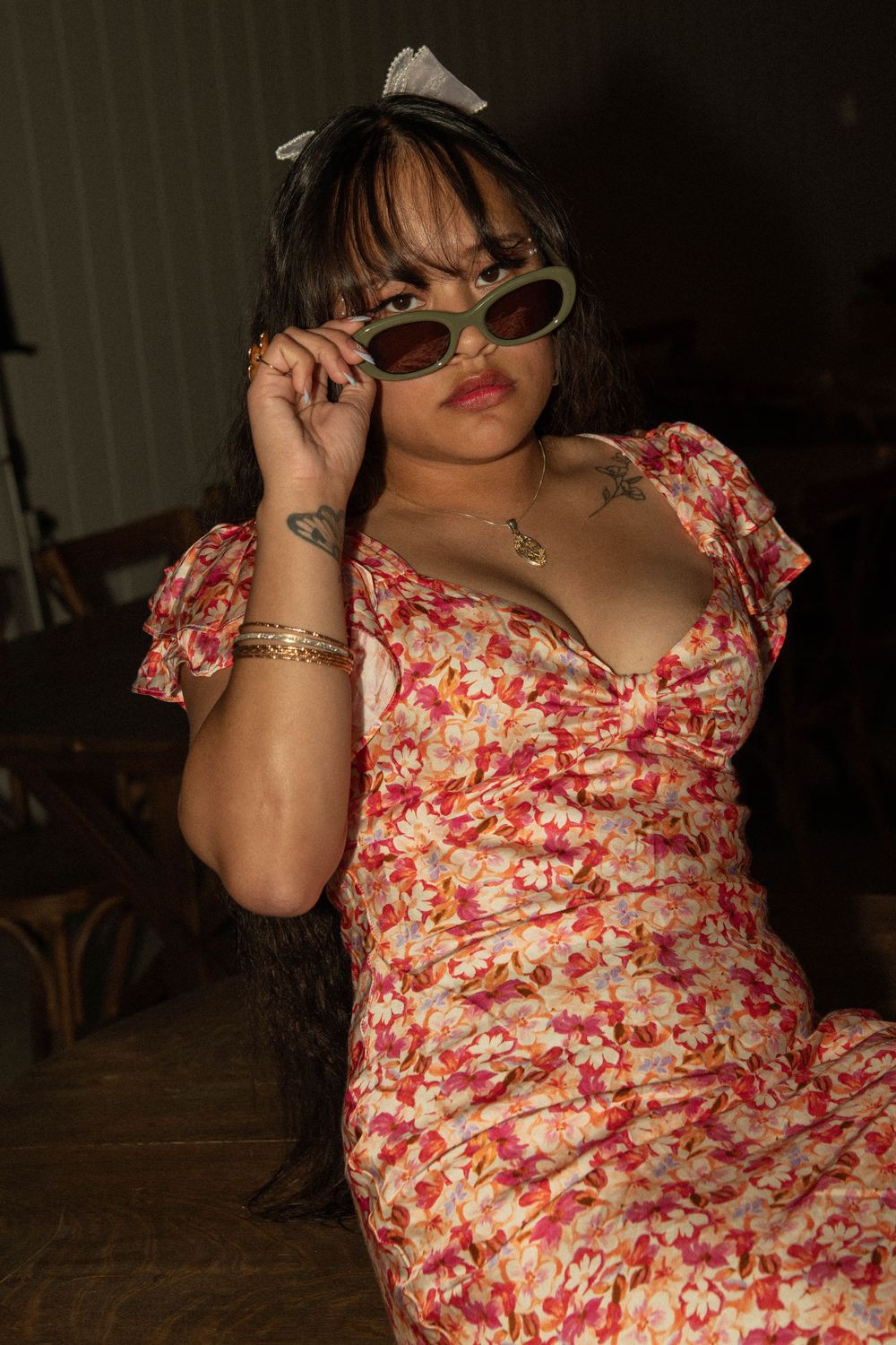 Woman in red floral dress posing with green cat-eye sunglasses in a dimly lit indoor setting.