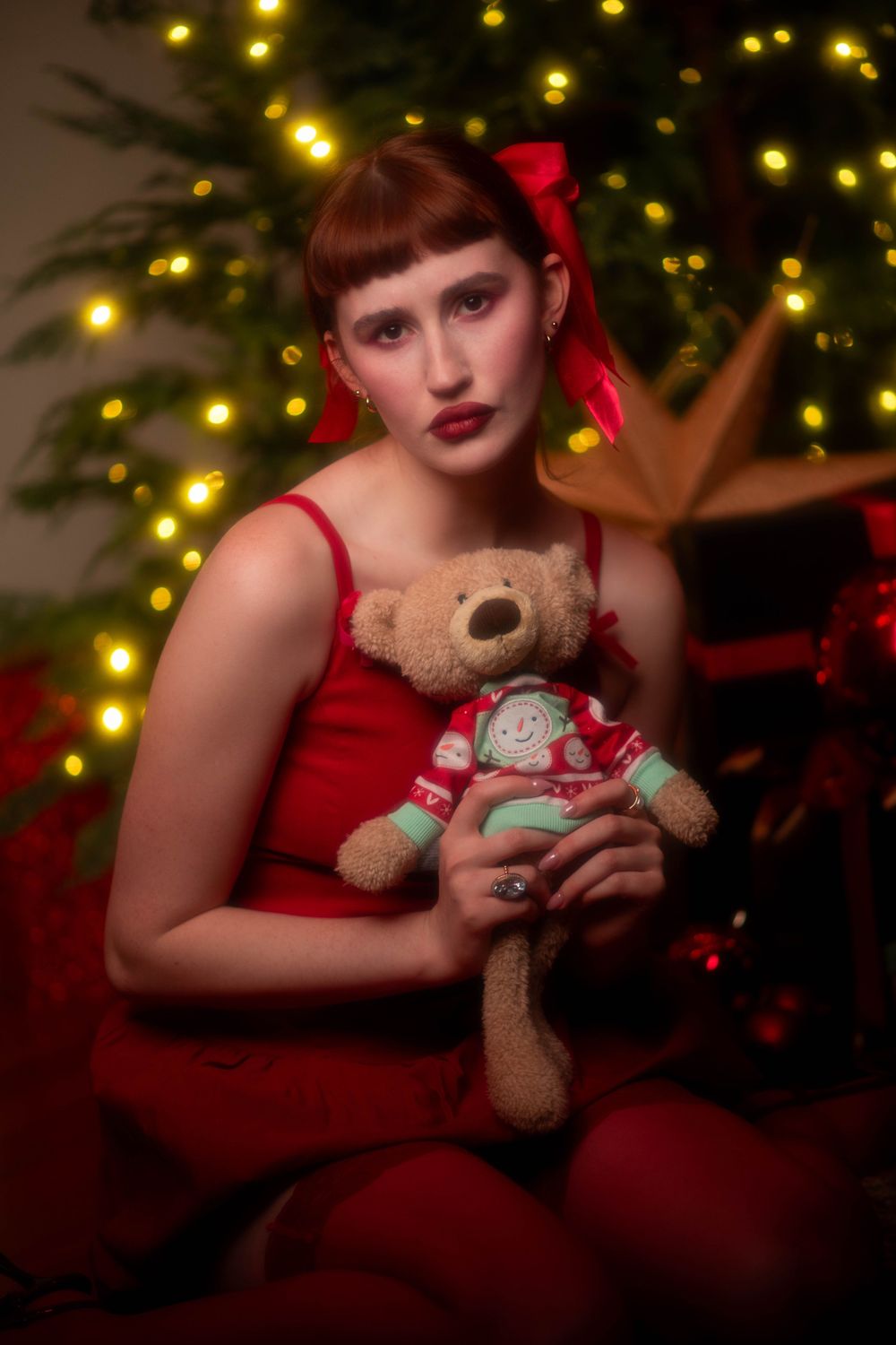 Young woman in red dress holding teddy bear in front of glowing Christmas tree with warm lights.