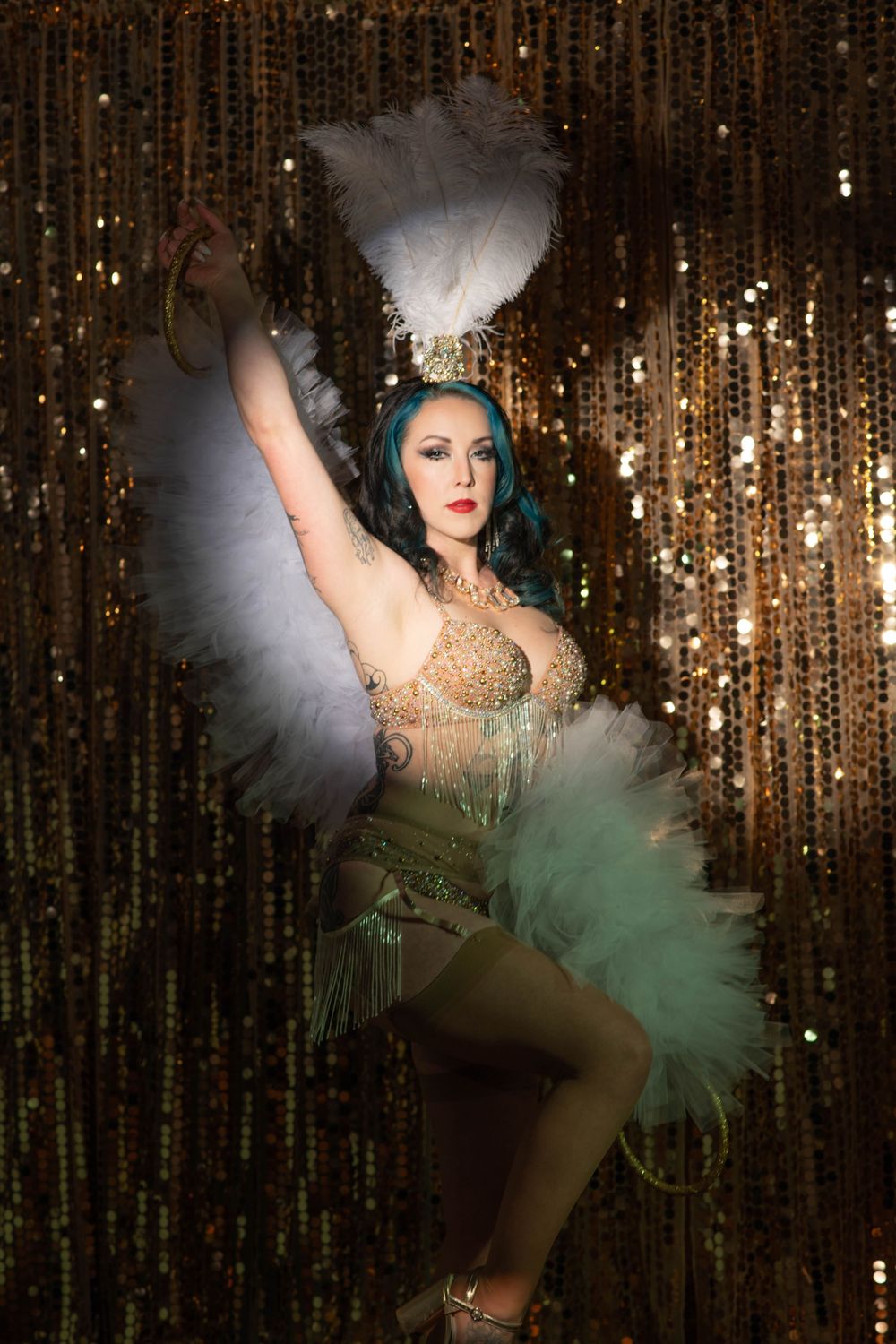 Burlesque dancer in gold and teal tutu with white feathers posing against gold sequin backdrop.