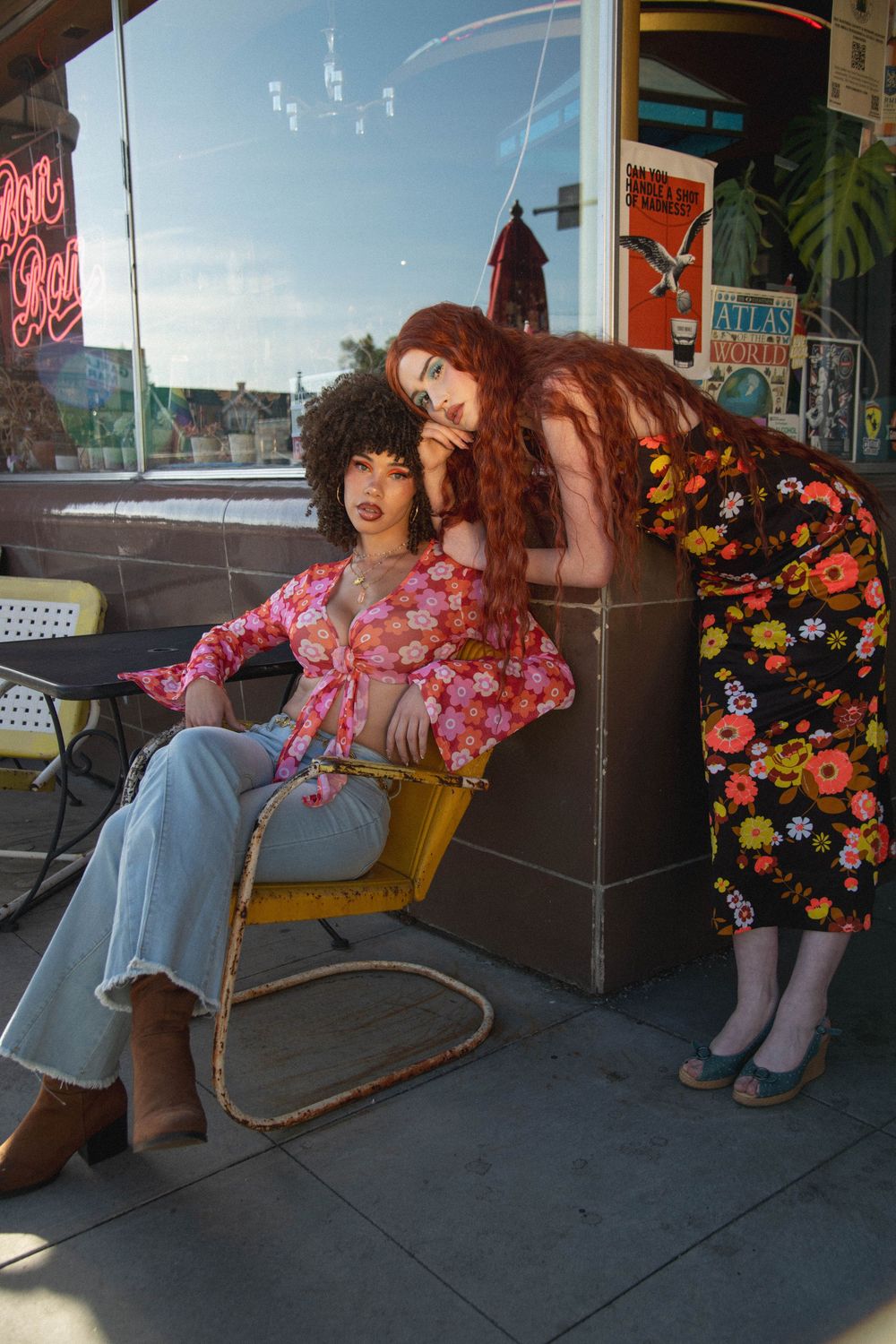 Two women in retro floral outfits pose outside a vintage shop, one seated in a yellow chair, the other leaning beside her.