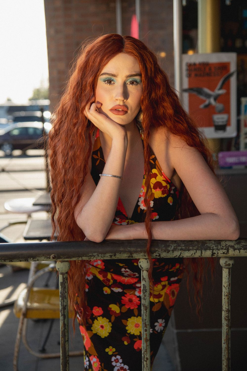 Young woman with long red wavy hair in floral dress leans on metal railing at outdoor cafe patio.