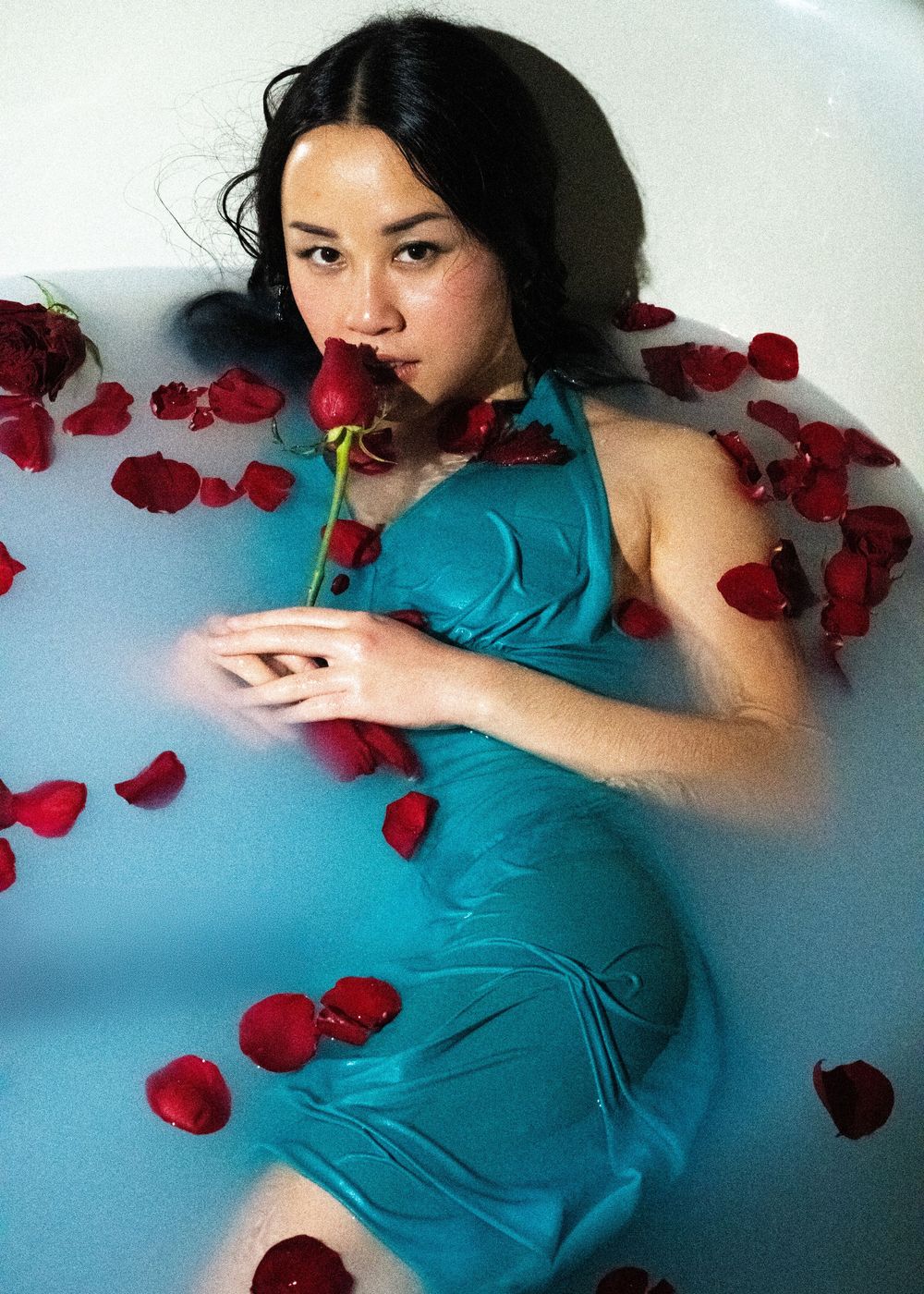 Woman in teal halter dress holds red rose lying in milky bath surrounded by rose petals.