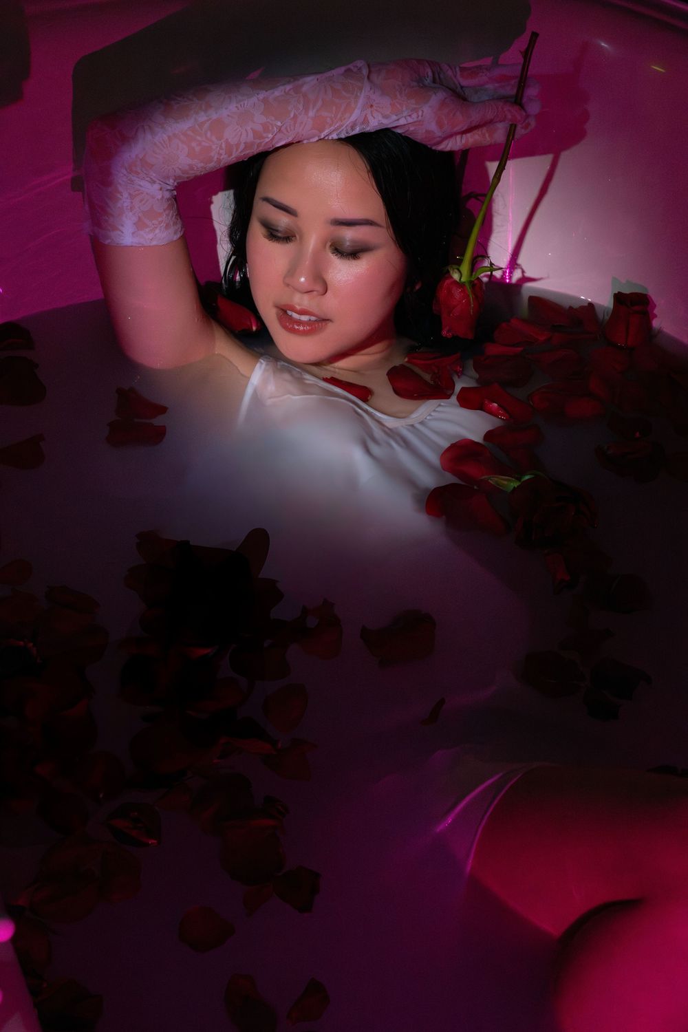 Woman lying in rose petal bath with pink lighting and lace gloves raised overhead.
