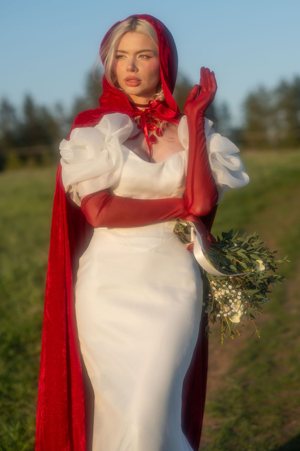 Blonde woman in red hooded cape and white bridal gown holds wildflower bouquet outdoors.