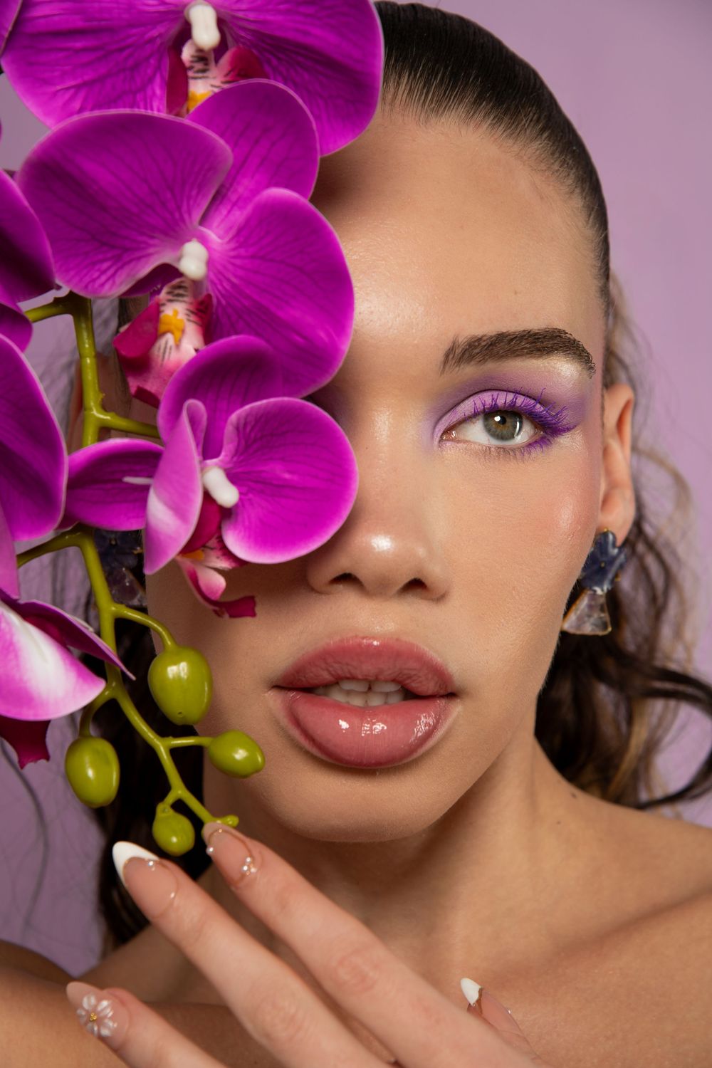 Close-up beauty shot of woman with purple eyeshadow holding pink orchid branch near face with French manicure.