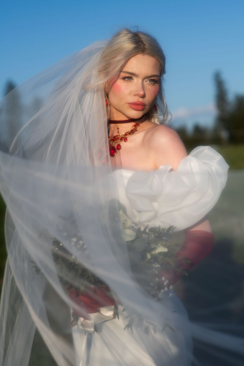 Blonde bride in white off-shoulder gown with red jewelry, veil flowing in wind against blue sky.