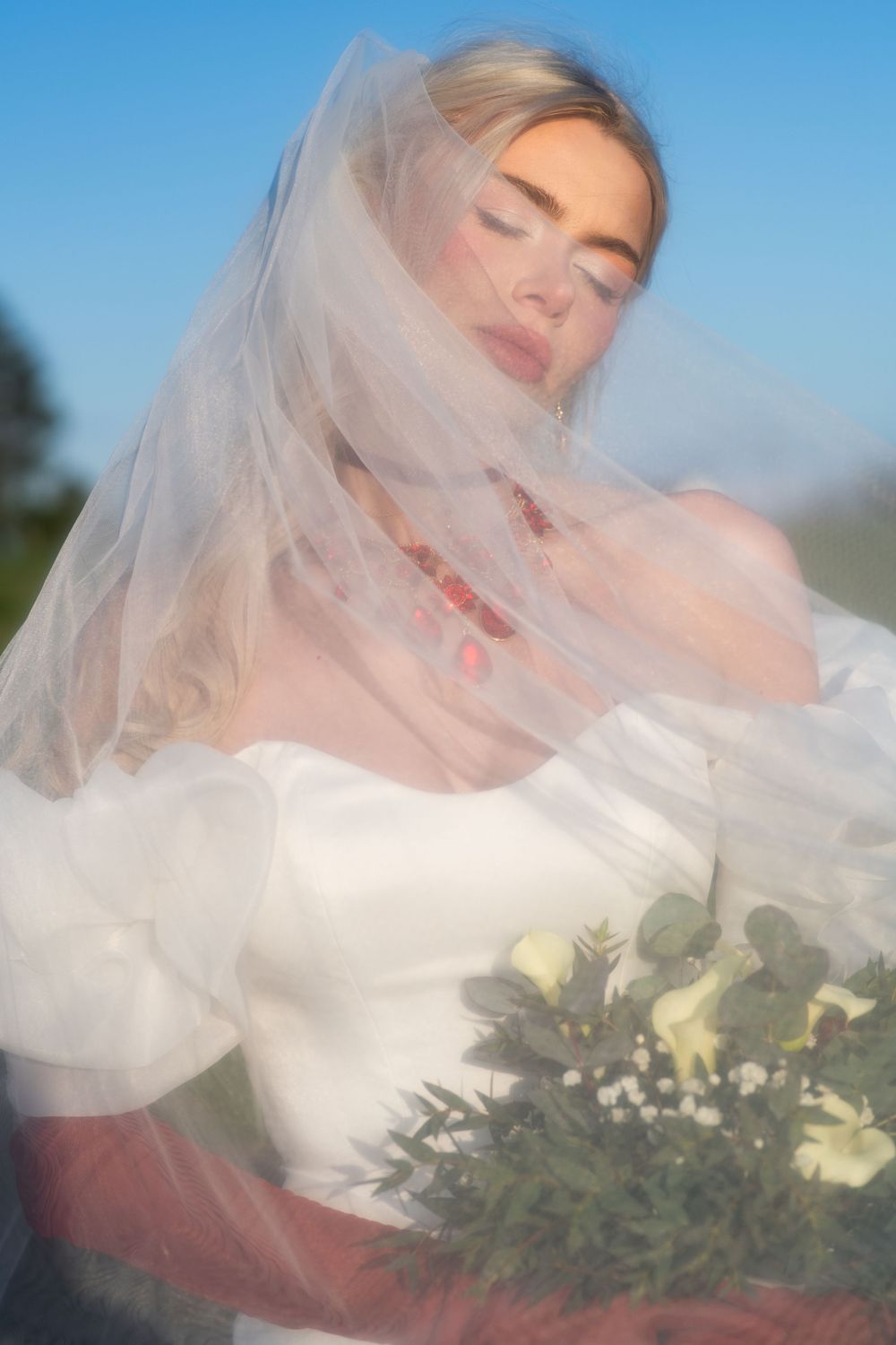 Bride with eyes closed, white veil draped over her, holding bouquet in white puff-sleeve gown.