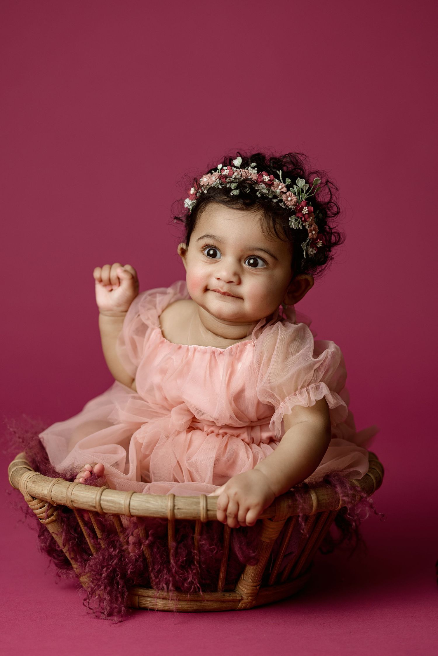 Baby wearing pink tulle dress and floral crown sits in wicker basket against burgundy backdrop.
