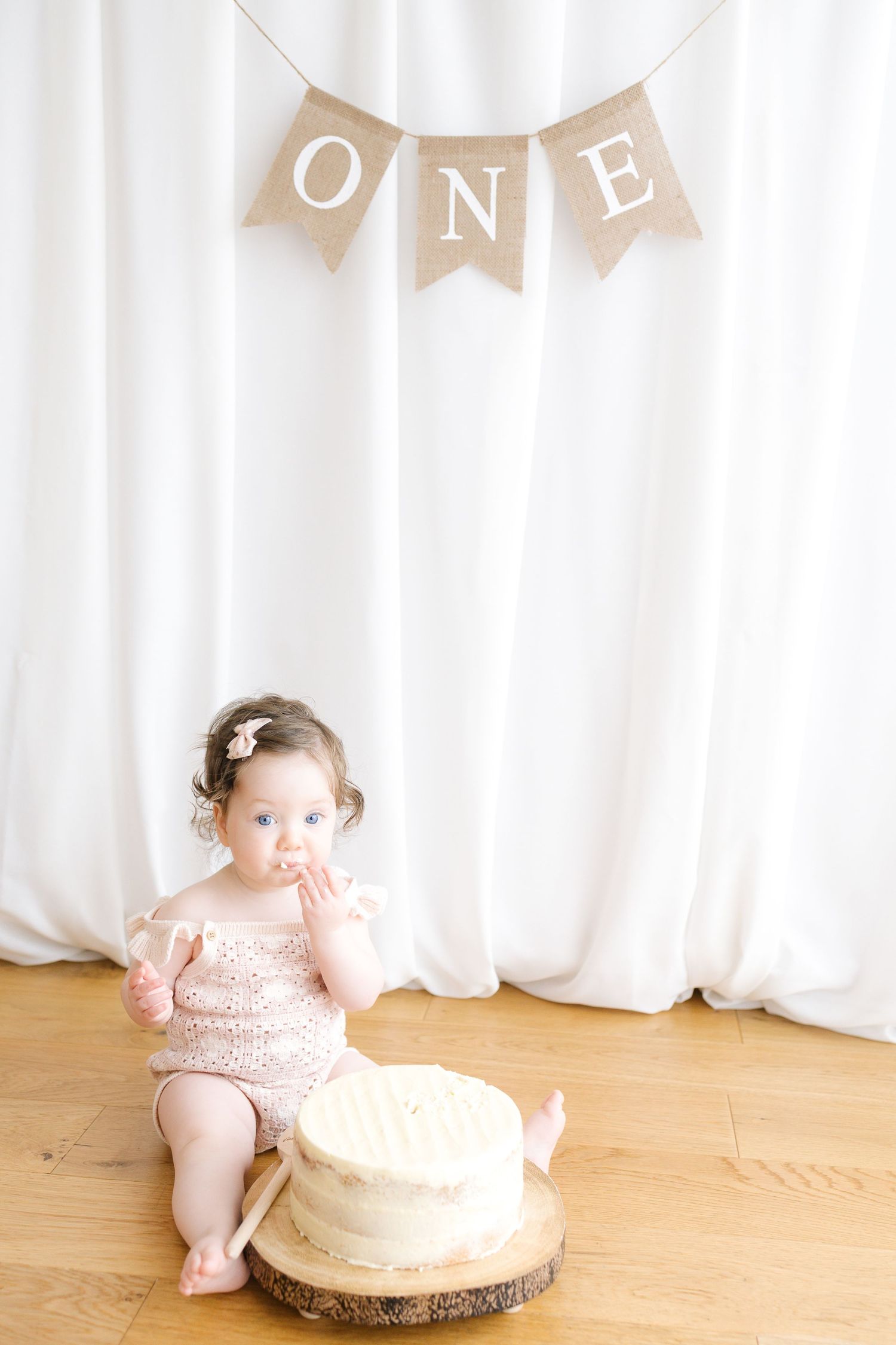 Baby in beige romper with teddy bear design next to birthday cake and ONE banner.