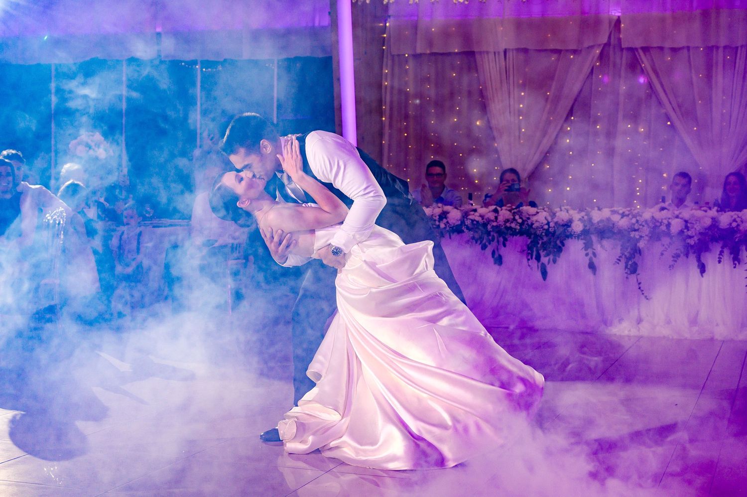 A couple performs their first dance at a wedding reception with dramatic purple lighting and fog effects on the dancefloor.