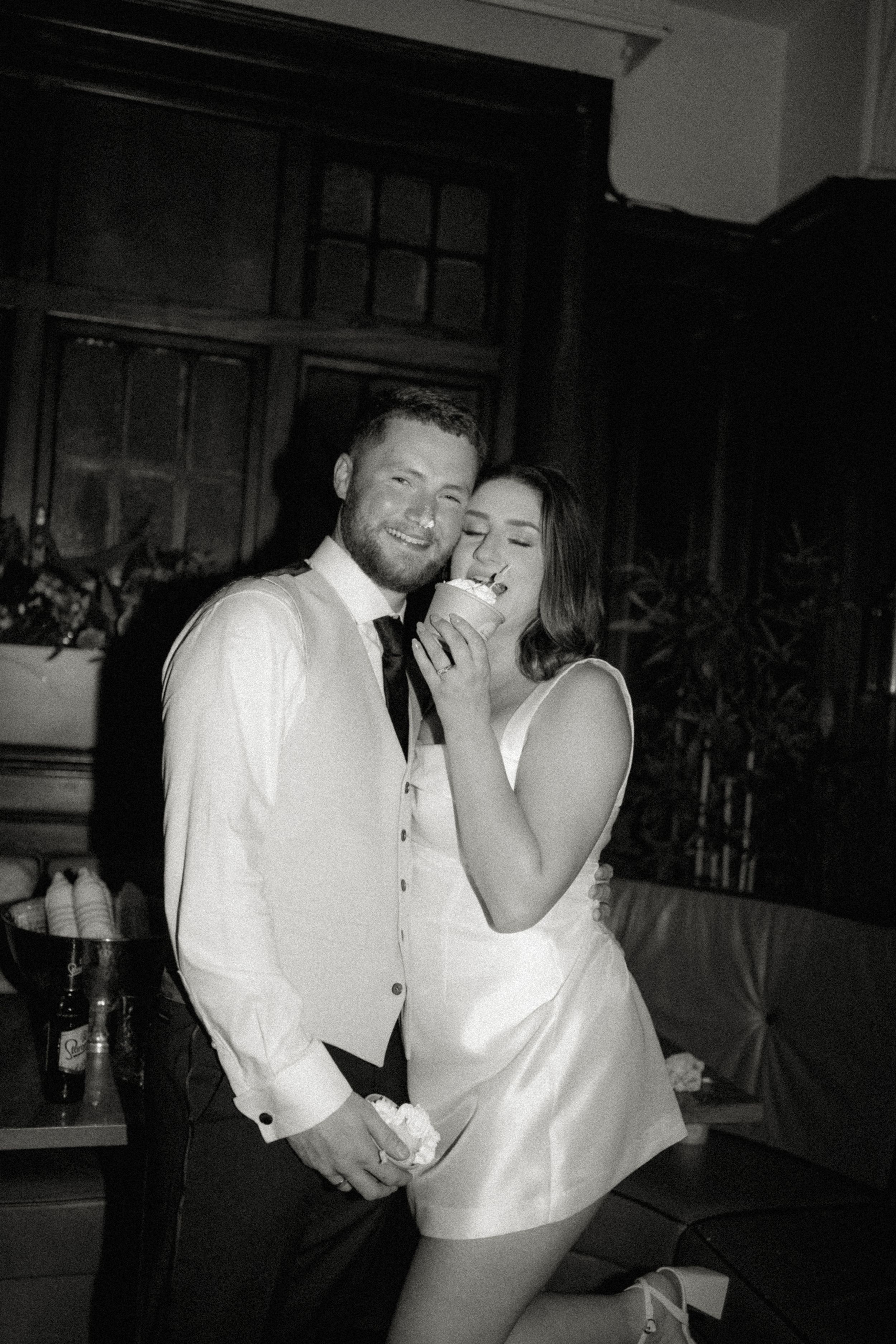 A candid black and white party photo showing a couple laughing and embracing in formal attire at an indoor venue.