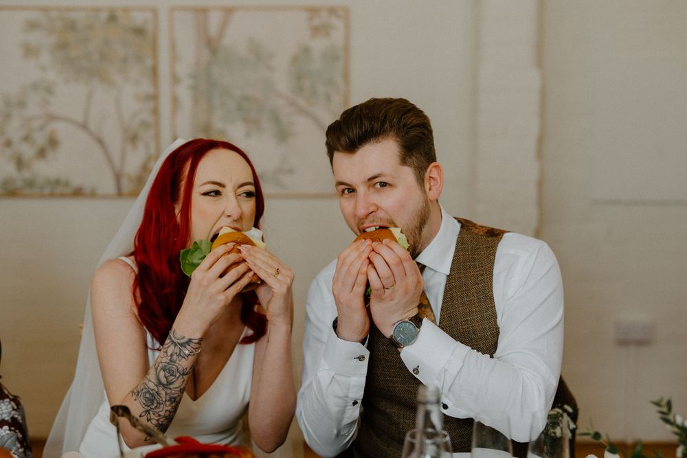 Two wedding guests in formal attire share a casual moment eating burgers at an elegant reception table.