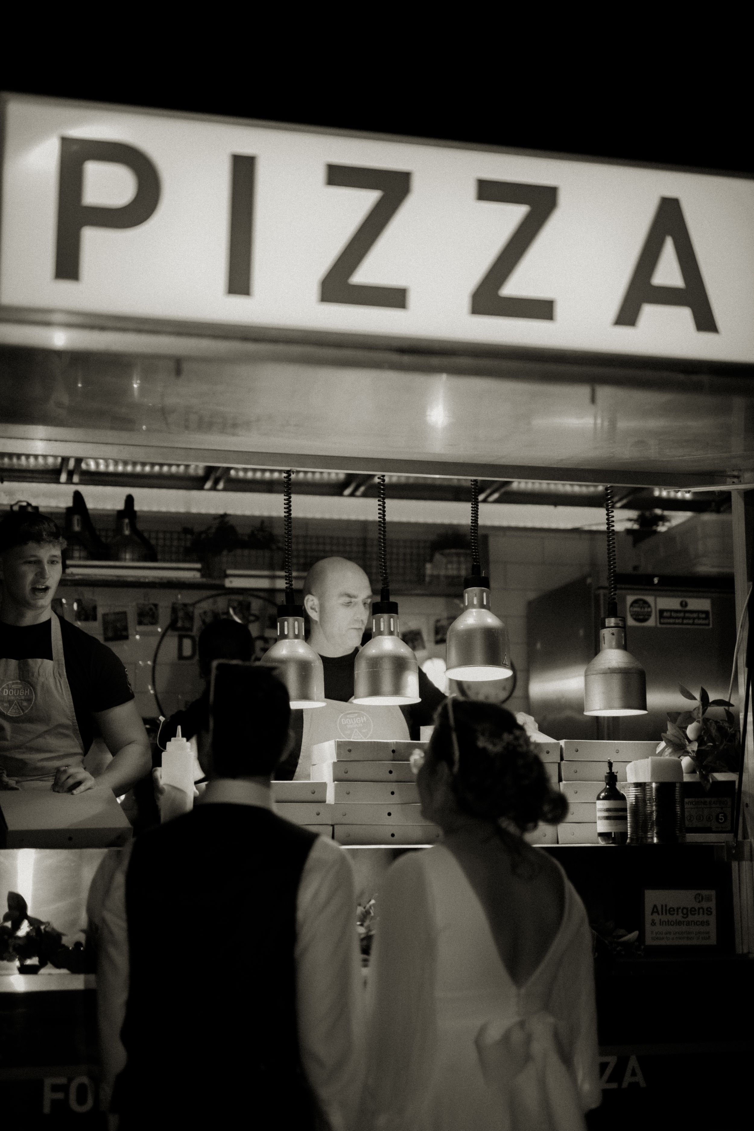 Black and white photo of a busy pizza counter with workers and stacked pizza boxes under a glowing PIZZA sign.