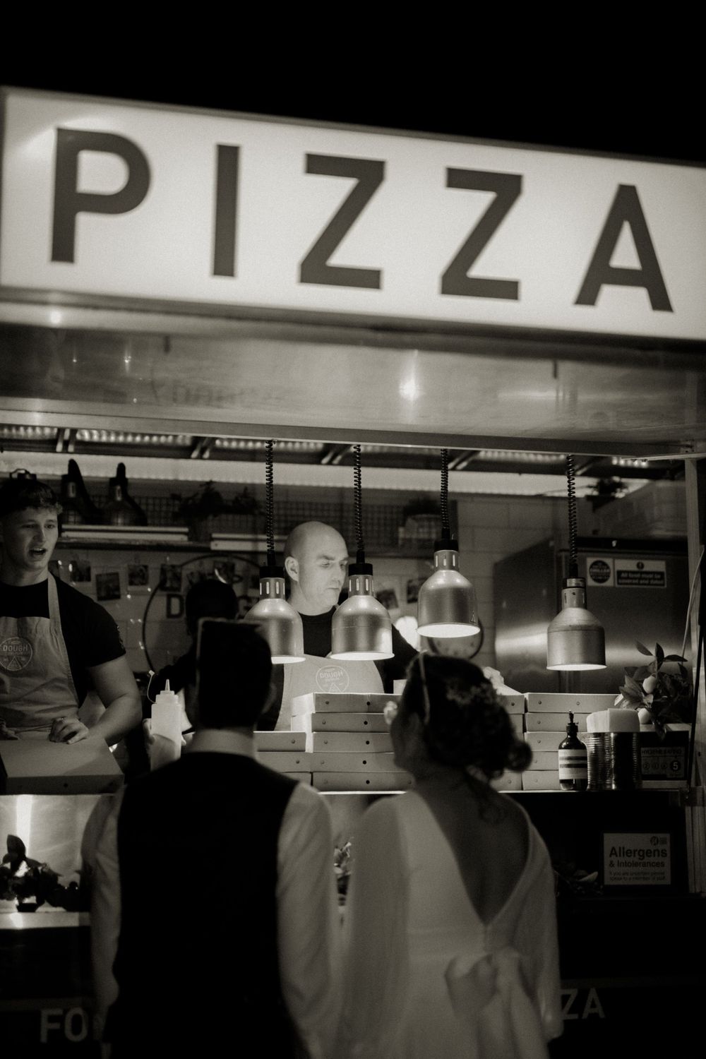 Black and white photo of a busy pizza counter with workers and stacked pizza boxes under a glowing PIZZA sign.