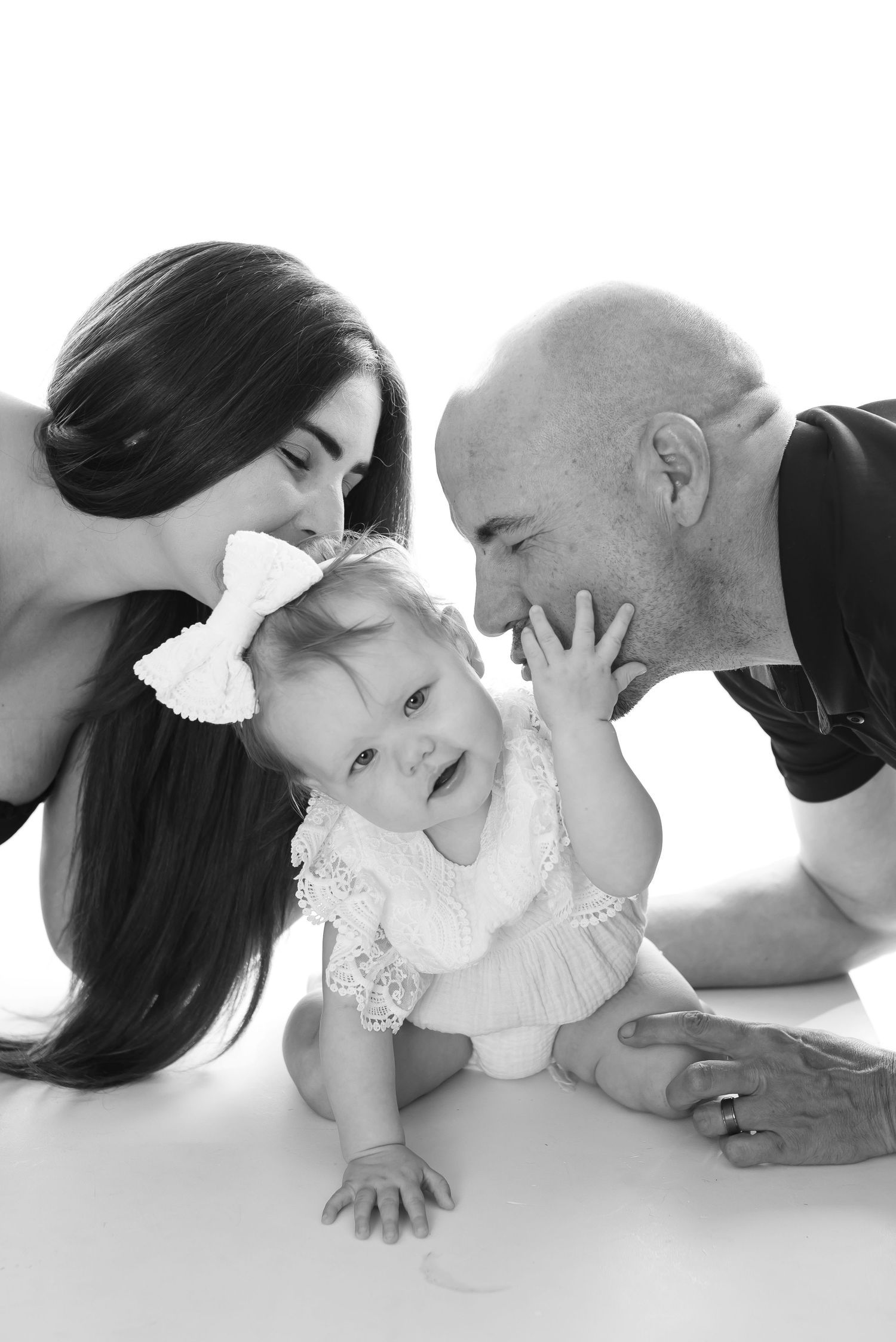 Black and white portrait shows a joyful family moment as a baby wearing a white bow dress plays while laying down.