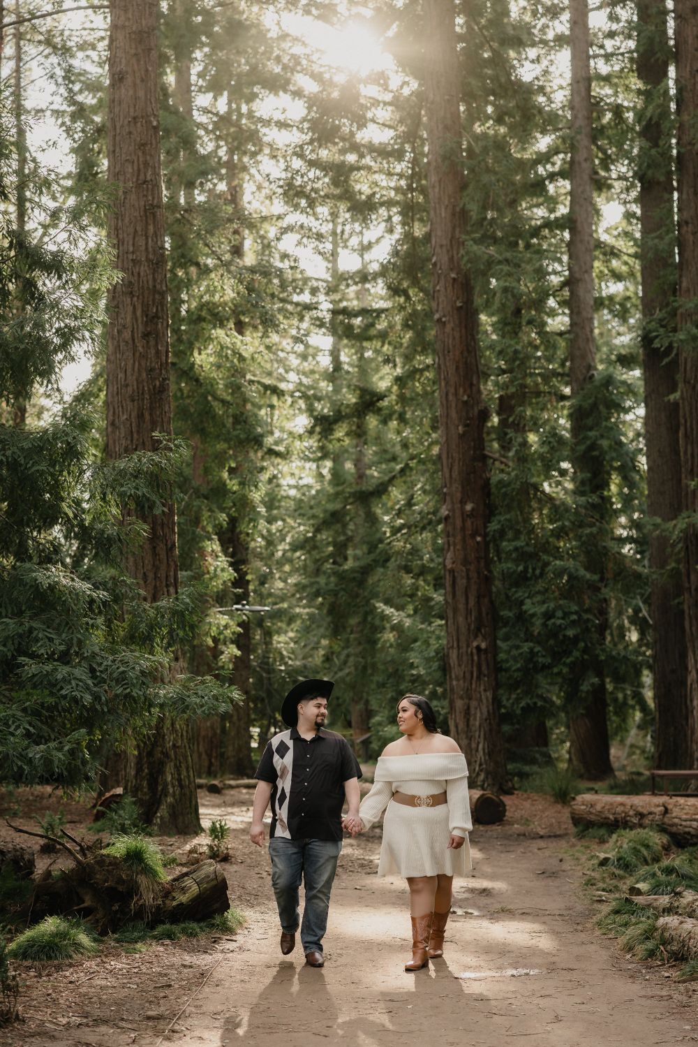 A couple strolling hand in hand down a dirt path through a majestic forest of tall pine trees.