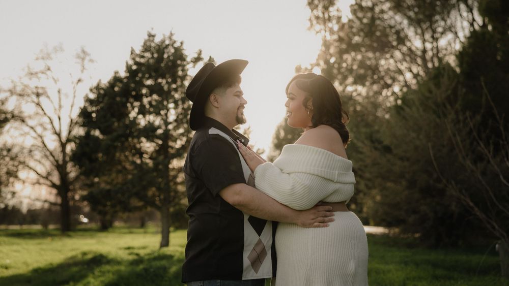Two people embrace in a romantic outdoor pose with soft sunlight filtering through trees in the background.