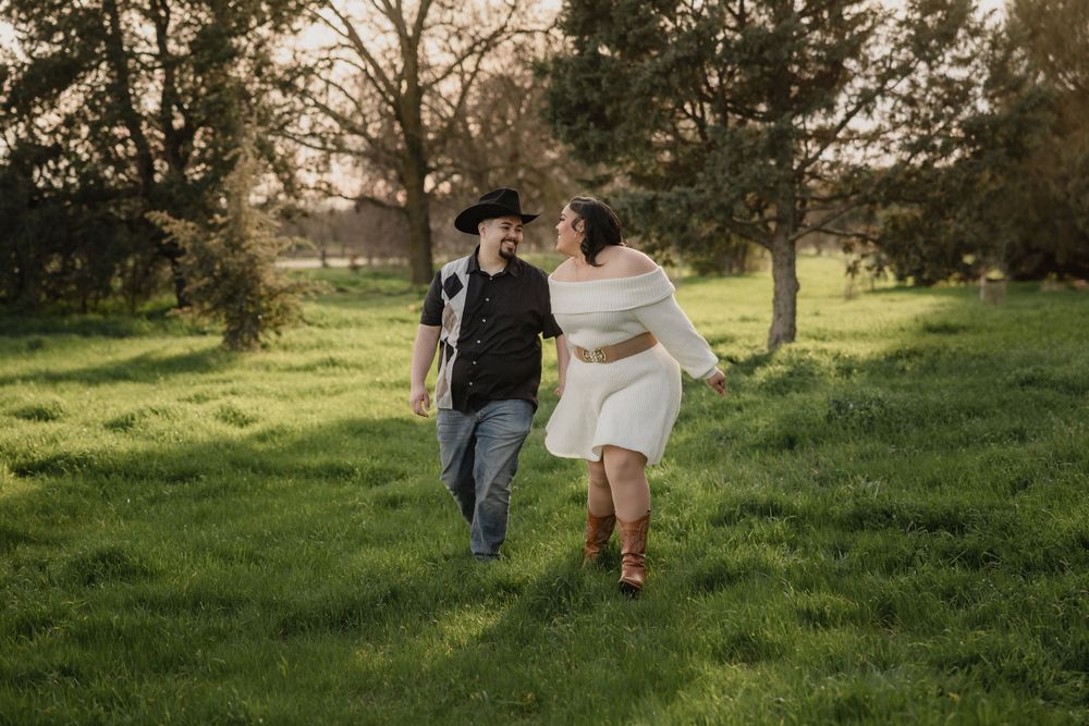 A joyful couple running through a sunlit grassy field with trees in the background.