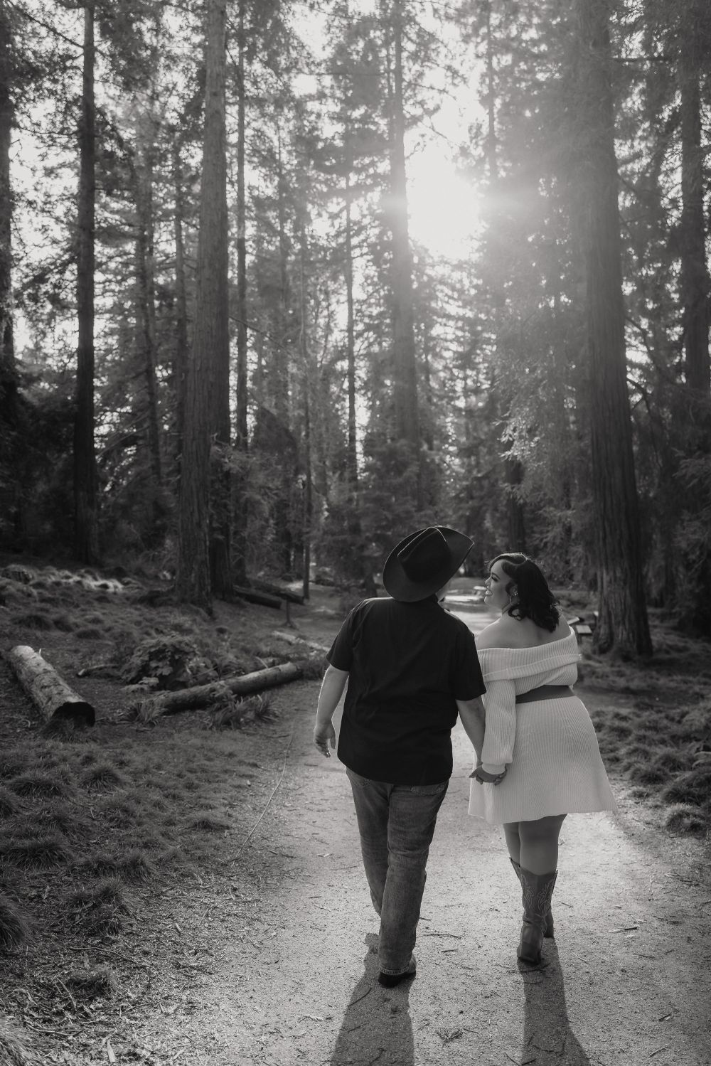 A couple walks hand in hand on a sunlit forest trail surrounded by tall trees in black and white.