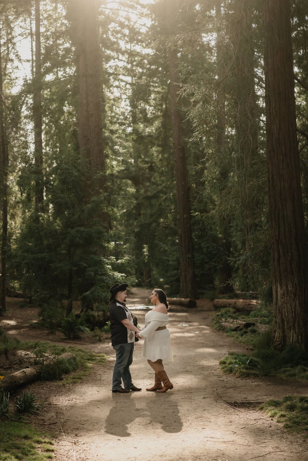 A couple stands together on a sunlit forest path surrounded by towering redwood trees in a romantic woodland setting.