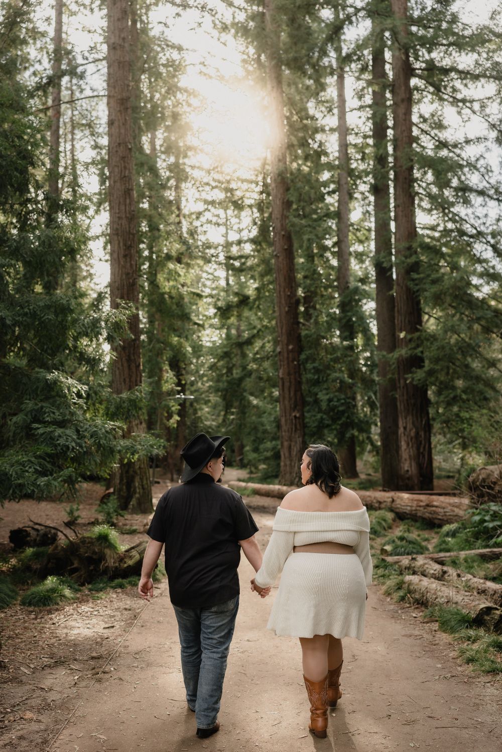 Two people walking down a sunlit forest path holding hands among towering evergreen trees.
