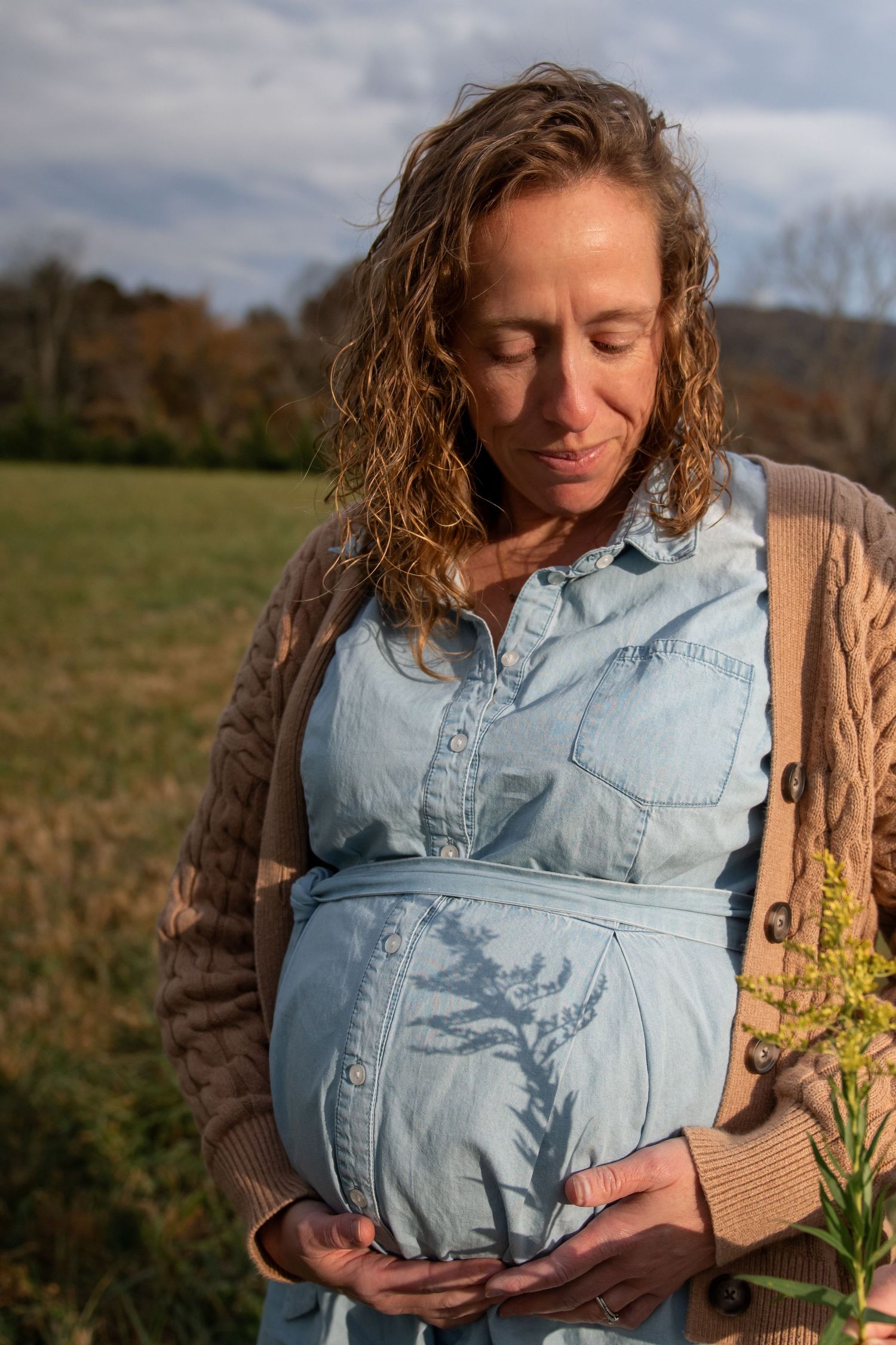 Person in light denim shirt stands in autumn field during golden hour with sunlight illuminating their profile.